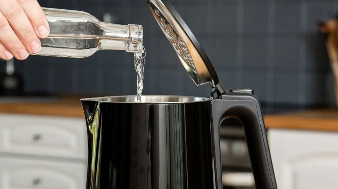 Main avec chiffon microfibre nettoyant l'intérieur d'une bouilloire programmable ouverte, bouteille de vinaigre blanc en arrière-plan sur plan de travail cuisine